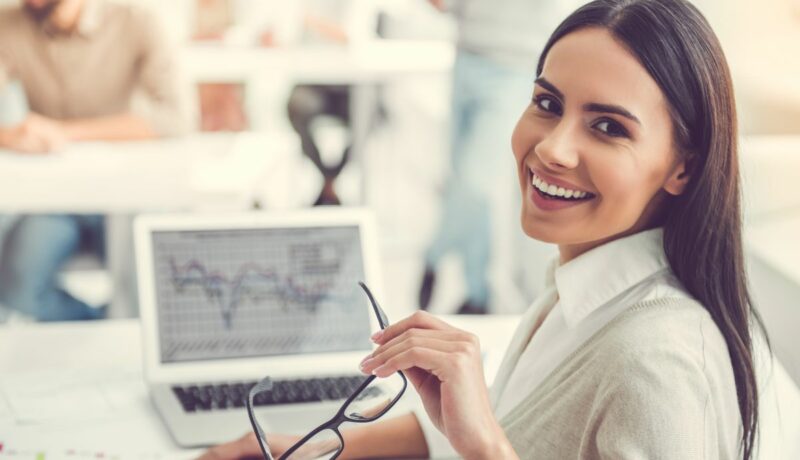 O femeie tânără, lucrează la birou, în fața calculatorului, se întoarce zâmbind, cu ochearii în mână