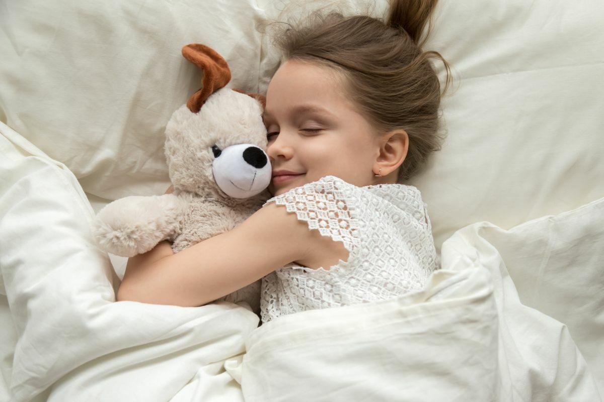 O fetiță, zâmbește, în timpul somnului, în vreme ce își strânge în brațe ursulețul