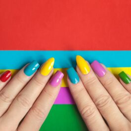 două mâini pe un fundal colorat cu unghiile făcute în nuanțe de roșu, albastru, galben, violet și verde care ilustrează cum să îți alegi forma ideală pentru unghii
