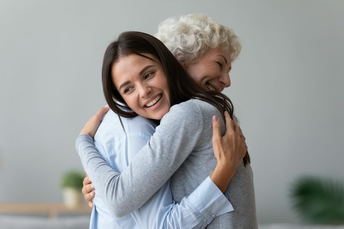 O femei tănără, îmbrățișează o alta în vârstă. Ambele zâmbesc și au capetele întoarse într-o parte
