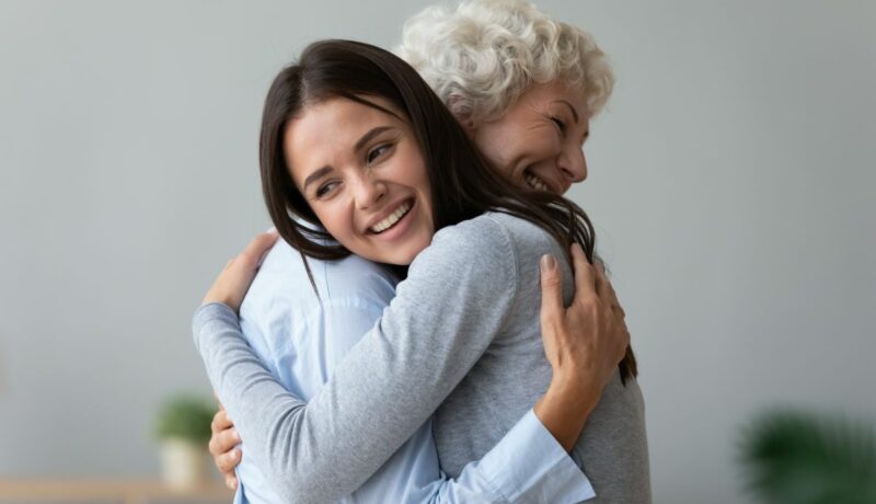 O femei tănără, îmbrățișează o alta în vârstă. Ambele zâmbesc și au capetele întoarse într-o parte