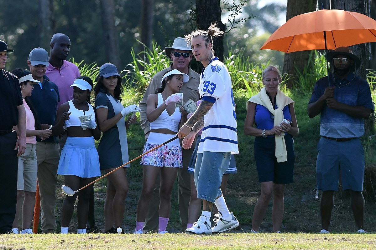 Justin Bieber îmbrăcat în pantaloni scurți albaștrii și un tricou alb larg în timp ce joacă golf alături de prietenii care îl susțin din spatele său