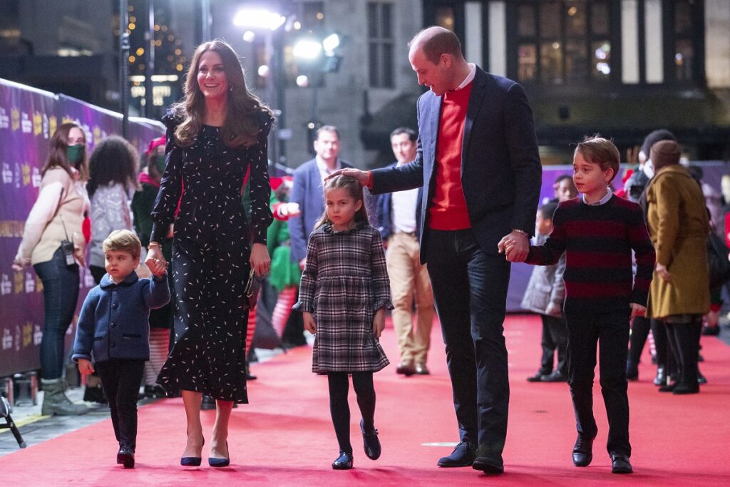 Kate Middleton într-o rochie elegantă neadră alături de Prințul William îmbrăcat la costum și o cămasă roșie, alături de cei trei copii pe covorul roșu care se îndreaptă către o excursie în care Kate Middleton a organizat o plimbare cu elicopterul pentru familia sa