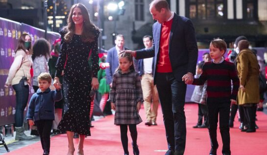 Kate Middleton într-o rochie elegantă neadră alături de Prințul William îmbrăcat la costum și o cămasă roșie, alături de cei trei copii pe covorul roșu care se îndreaptă către o excursie în care Kate Middleton a organizat o plimbare cu elicopterul pentru familia sa