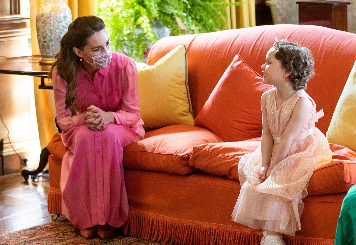 Kate Middleton într-o rochie lungă roz stând pe o canapea orange alături de micuța Mila Sneddon îmbrăcată la fel ca o prințesă