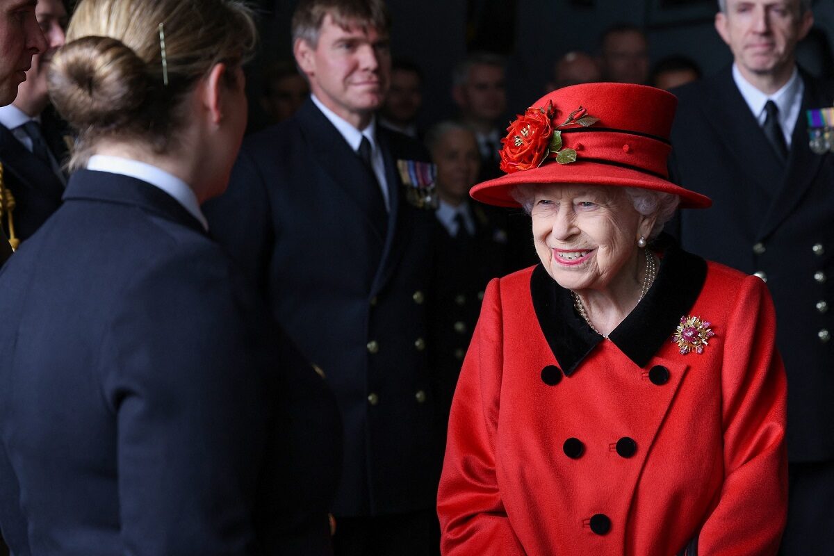 Regina Elisabeta într-un costum roșu la vizita portavionului HMS Regina Elisabeta în timp ce se salută cu oficialitățile de la bord și poartă în piept broșa primită de la Printul Philip cadou în 1966