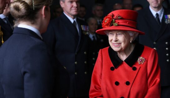 Regina Elisabeta într-un costum roșu la vizita portavionului HMS Regina Elisabeta în timp ce se salută cu oficialitățile de la bord și poartă în piept broșa primită de la Printul Philip cadou în 1966