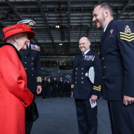 Regina Elisabeta într-un costum roșu în timp ce se salută cu căpitanul forțelor marine Angus Essenhigh în timpul vizitei de la bordul portavionului HMS Regina Elisabeta