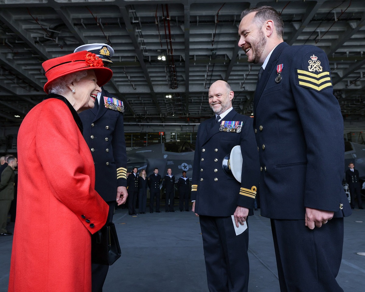 Regina Elisabeta într-un costum roșu în timp ce se salută cu căpitanul forțelor marine Angus Essenhigh în timpul vizitei de la bordul portavionului HMS Regina Elisabeta