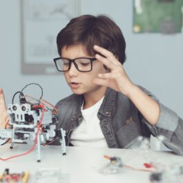 Un băiat construiește un robot, în timp ce are o cămașă gri și ochelari și stă la o masă, iar în fața sa se află roboțelul
