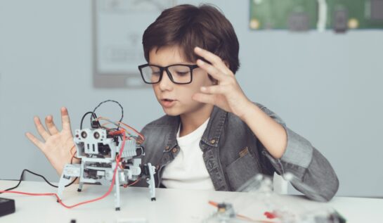 Un băiat construiește un robot, în timp ce are o cămașă gri și ochelari și stă la o masă, iar în fața sa se află roboțelul
