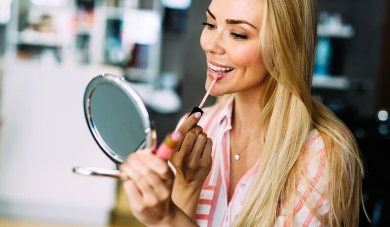O femeie blondă, ține o oglindă rotundă în mână și își aplică un ruj pe buze