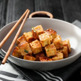 Într-un bol alb sunt cuburi de tofu prăjit. Pe farfurie sunt niște bețișoare pentru servit.
