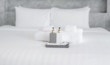 un pat cu așternuturi albe pe care sunt așezate două sticluțe de culori alb și gri, alături de prosoape albe pentru a demonstra de ce sunt folosite așternuturile albe în camerele de hotel