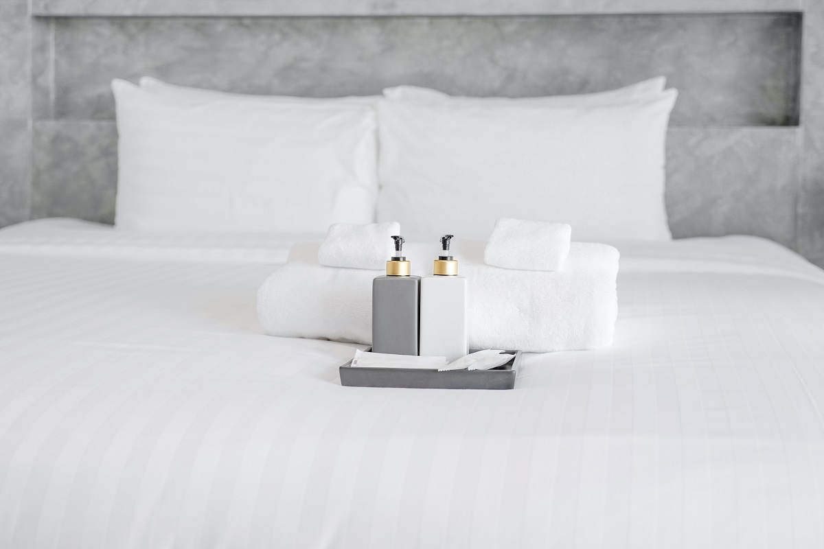 un pat cu așternuturi albe pe care sunt așezate două sticluțe de culori alb și gri, alături de prosoape albe pentru a demonstra de ce sunt folosite așternuturile albe în camerele de hotel
