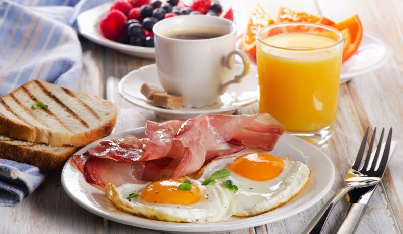 Pe masă este o farfurie cu două ouă ochiuri, cu niște felii de bacon. Alături se află o cafea și un suc de portocale și câteva fructe: zmeură, portocale și afine