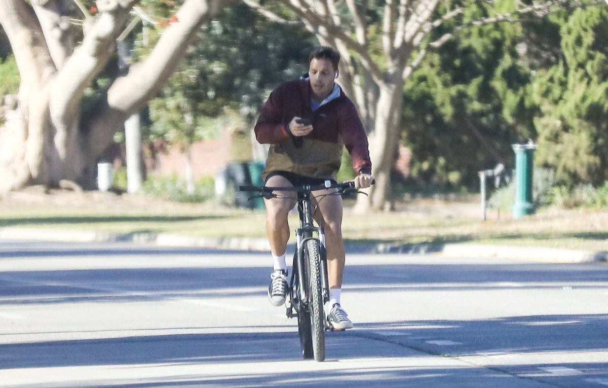 Joseph Baena se plimbă cu bicicleta și își verifică telefonul. Poartă o geacă groasă și pantaloni scurți