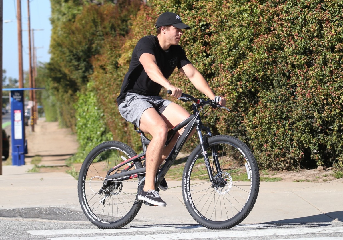 Joseph Baena se plimbă cu bicicleta. Poartă o șapcă neagră, un tricou negru, pantaloni scurți gri și pantofi sport negri.