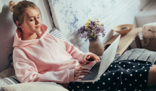 O femeie care stă în pat cu un laptop în brațe, în timp ce are părul strâns în coc, poartă un hanorac roz și pantaloni negri cu buline albe