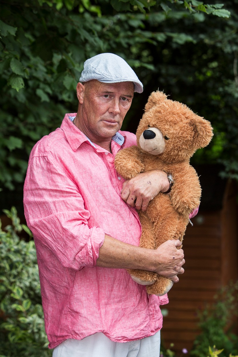 Mark Evans, într-o imagine în care ține un urs de pluș în brațe, îmbrăcat cu o cămașă roz, pantaloni deschiși la culoare și șapcă