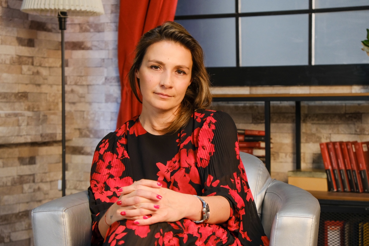 Olga Abramescu, fotografiată cu mâinile împreunate peste picior, într-o rochie neagră cu imprimeu floral, la interviul pentru CaTine.ro