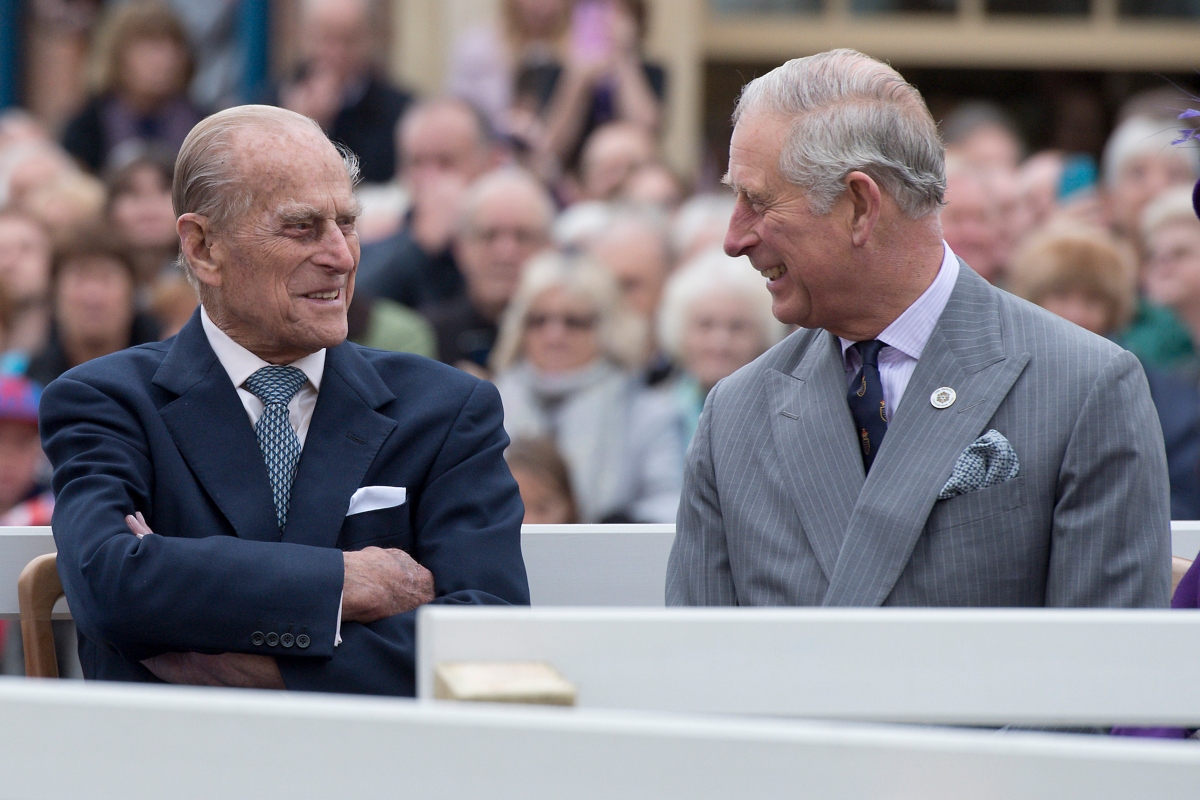 În imagine sunt Prințul Charles cu Prințul Philip. Ambii poartă costume elegante, de culori închise.