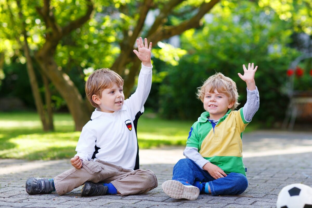 Doi copii stau așezați pe asfalt și țin câte o mână ridicată