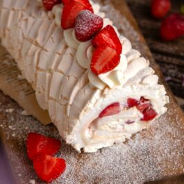 O ruladă de bezea cu cremă de vanilie și căpșuni, așezată pe un blat pudrat cu zahăr pubdră, alături de căpșuni.