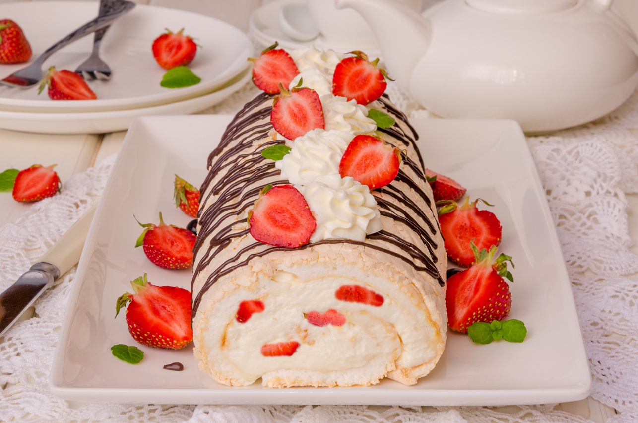 Ruladă de bezea pe un platou alb, decorată cu ciocolată și căpșuni
