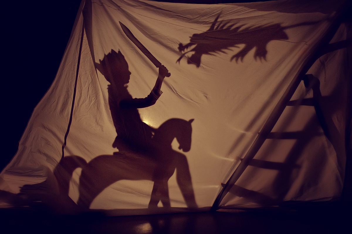 O piesă de teatru în umbră unde se poate observa un copil călare pe un cal în timp ce se luptă cu un dragpn în spatele unui așternut alb luminat cu o lampă pentru a forma umbre