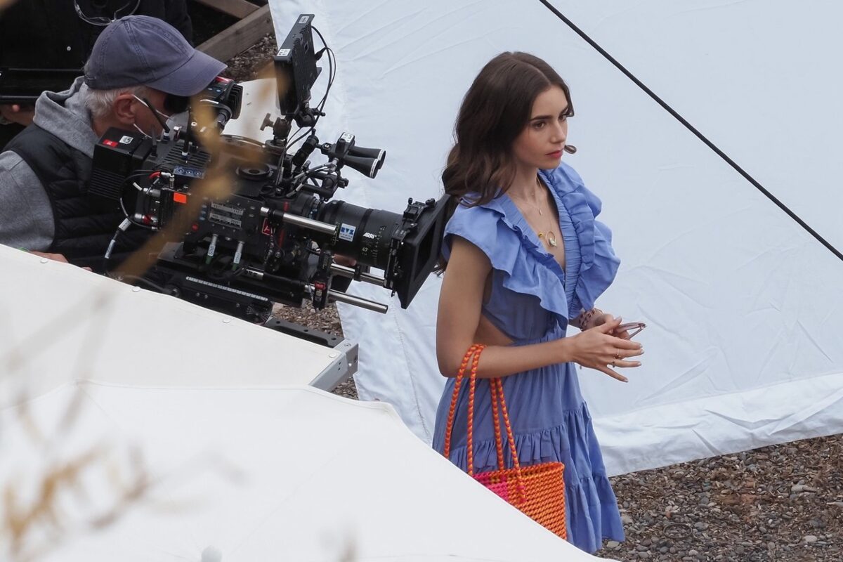Un cadru din Emily în Paris, sezonul 2. Lily Collins poartă o rochie albastră și în spate are un cameraman care filmează.