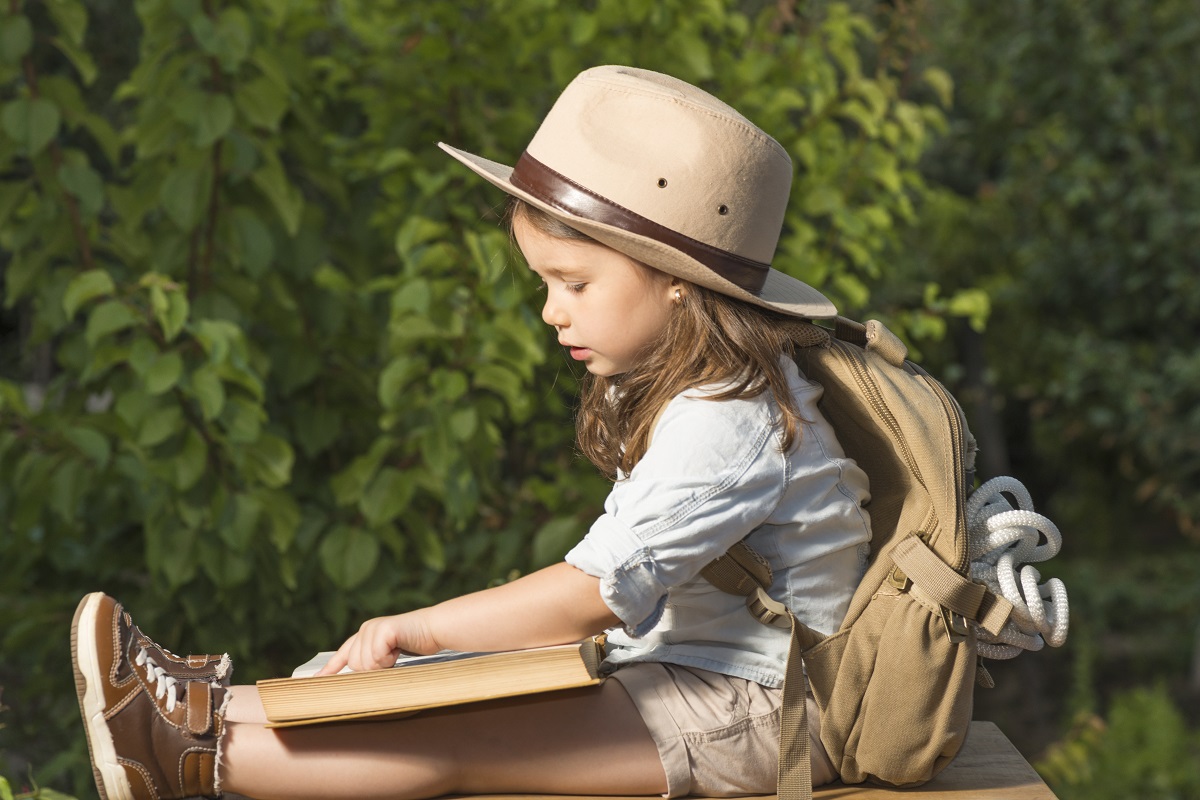 O fetiță îmbrăcată pentru o călătorie cu pălărie, rucsac de munte și pantofi speciali în timp ce stă jos și citește dintr-o carte pentru a răspunde la întrebările vânătoarei de comori, una din cele cinci activități creative de 1 iunie