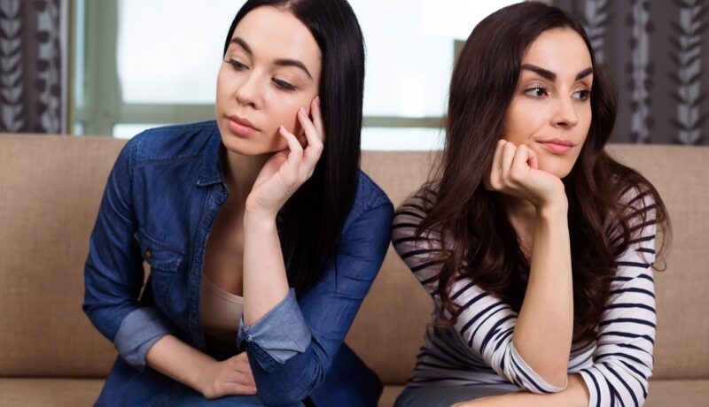 Două femei care stau pe o canapea în timp ce privesc în direcții diferite și sunt supărate una pe cealaltă din cauza unor semne de gelozie din relația lor de prietenie