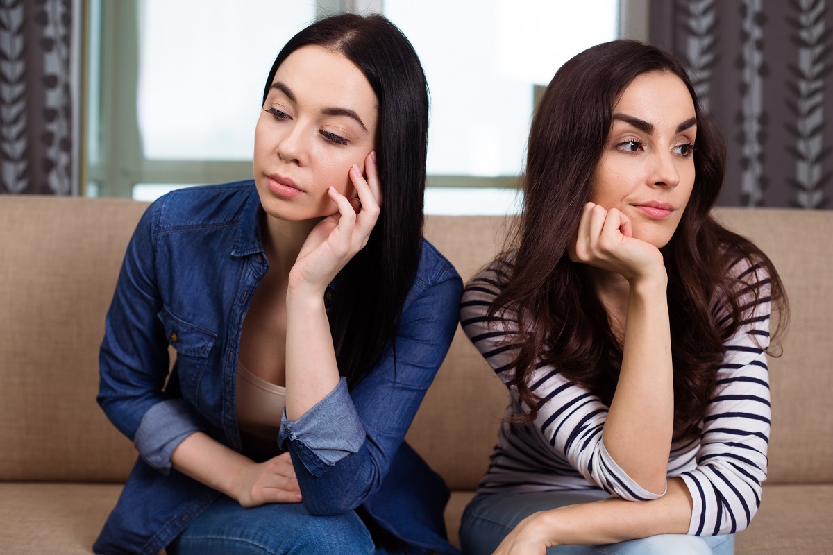 Două femei care stau pe o canapea în timp ce privesc în direcții diferite și sunt supărate una pe cealaltă din cauza unor semne de gelozie din relația lor de prietenie