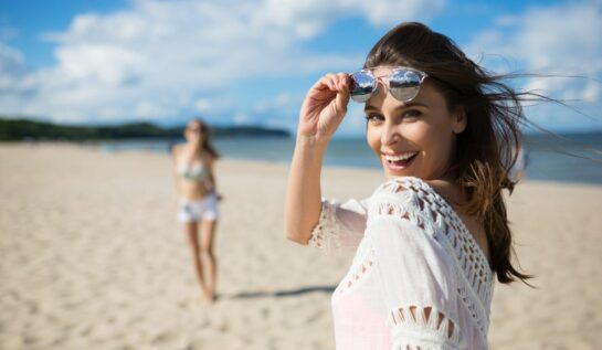 O femeie frumoasă, îmbrăcată în alb, își ridică ochelarii de soare, în timp ce se află pe plajă