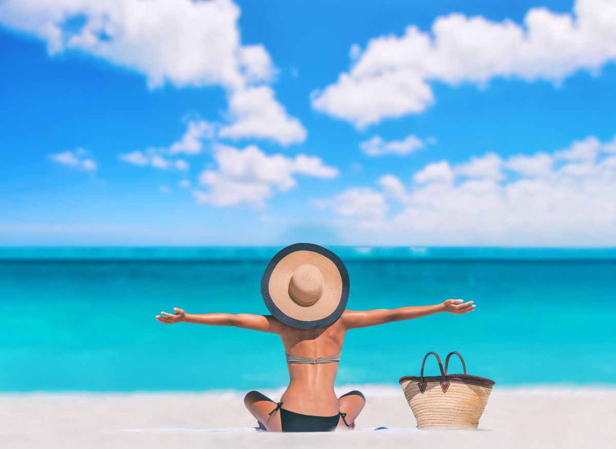 O femeie stă pe plajă, cu fața spre mare și spatele spre camera. Are o pălărie mare de paie și își întinde brațele