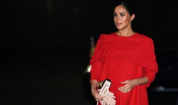 Meghan Markle într-o rochie roșie în timp ce ăși ține mâna pe burtică este una dintre cele trei femei din Familia Regală a Marii Britanii care au devenit mame după vârsta de 30 de ani
