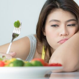 O femeie care ține cotul pe masă în timp ce privește o farfurie cu legume și este indecisă dacă să le consume deaorece imaginea sa de sine este scăzută și suferă de o tulburare de alimentație