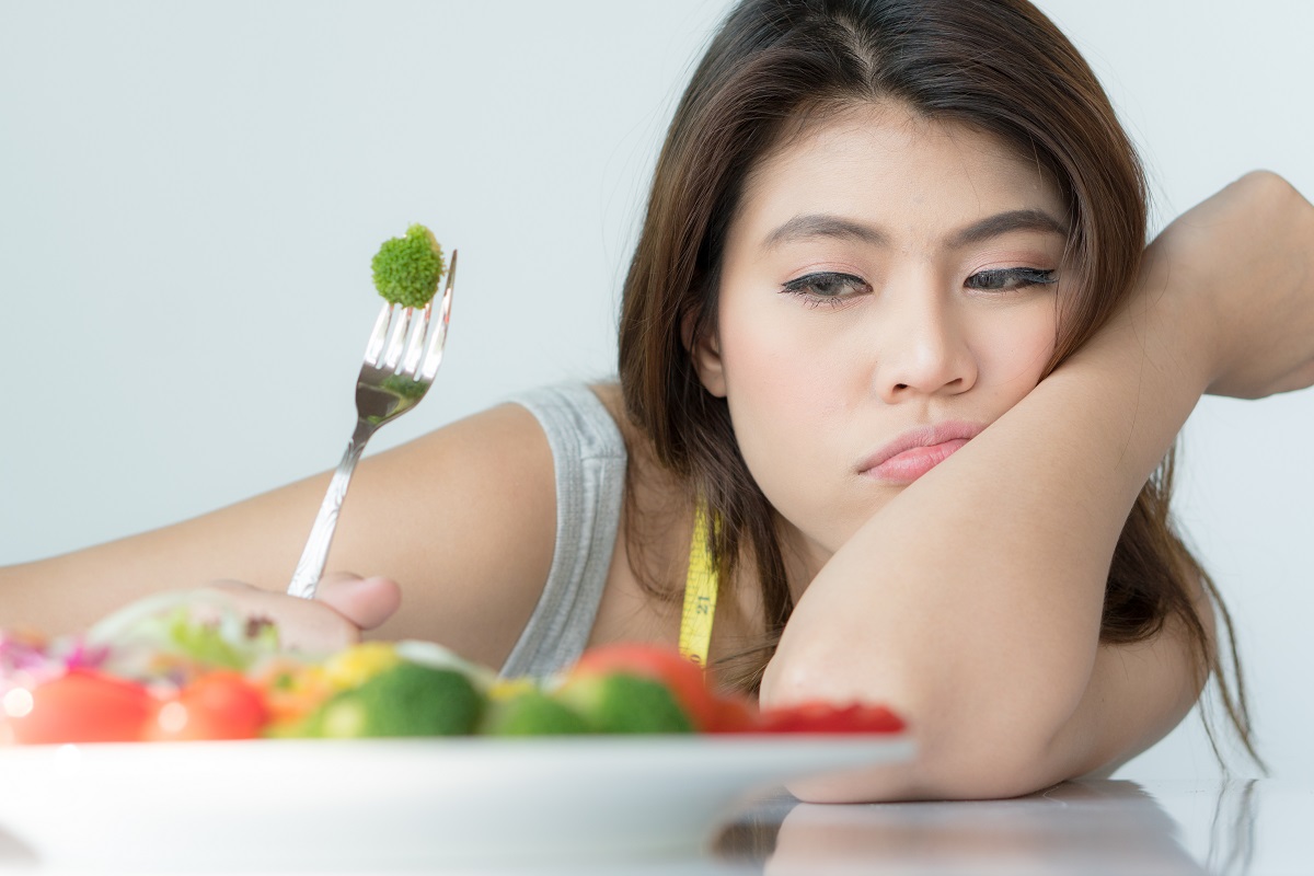 O femeie care ține cotul pe masă în timp ce privește o farfurie cu legume și este indecisă dacă să le consume deaorece imaginea sa de sine este scăzută și suferă de o tulburare de alimentație