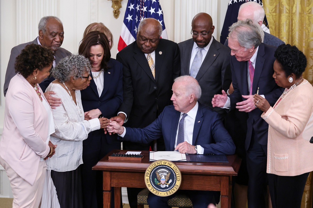 Președintele Joe Biden înconjurat de Opal Lee și membrii ai senatului în timp ce îi oferă stiloul activistei Opal Lee