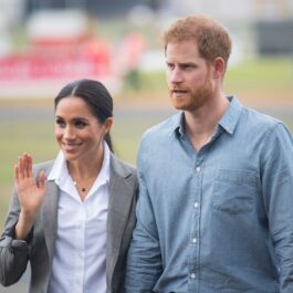 Meghan Markle, care nu va participa la dezvelirea statuii Prințesei Diana, îmbrăcată într-o cămașă albă și un sacou gri în timp ce face cu mâna oamenilor și este alături de soșțul său, Prințul Harry îmbrăcat într-o cămașă de culoare albastră