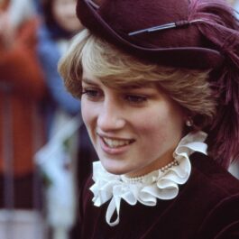 Portret cu Lady Di în timp ce zâmbește oamenilor și poartă o cămasă violet cu guler bufant alb, la care a accesorizat o pălărie mov, într-unul din acele momente emblematice ale Prințesei Diana