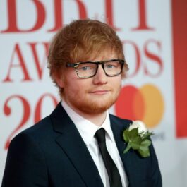 Ed Sheeran poartă un costum negru și o cămașă albă, cu un trandafir alb la rever
