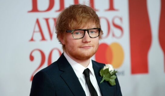 Ed Sheeran poartă un costum negru și o cămașă albă, cu un trandafir alb la rever