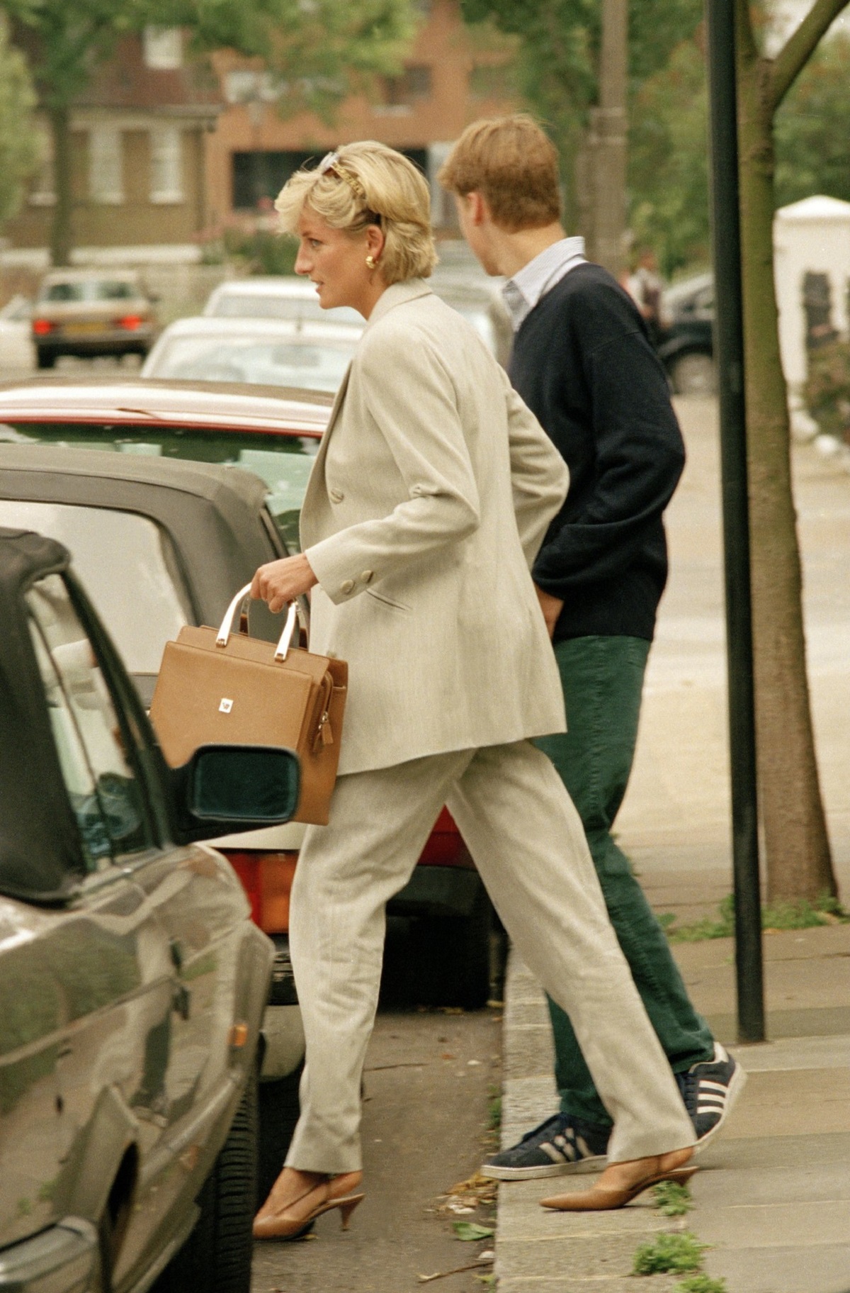 Prințesa Diana într-un costum gri în timp ce ține în mână o poșetă și îl conduce pe fiul său, Prințul William la mașină
