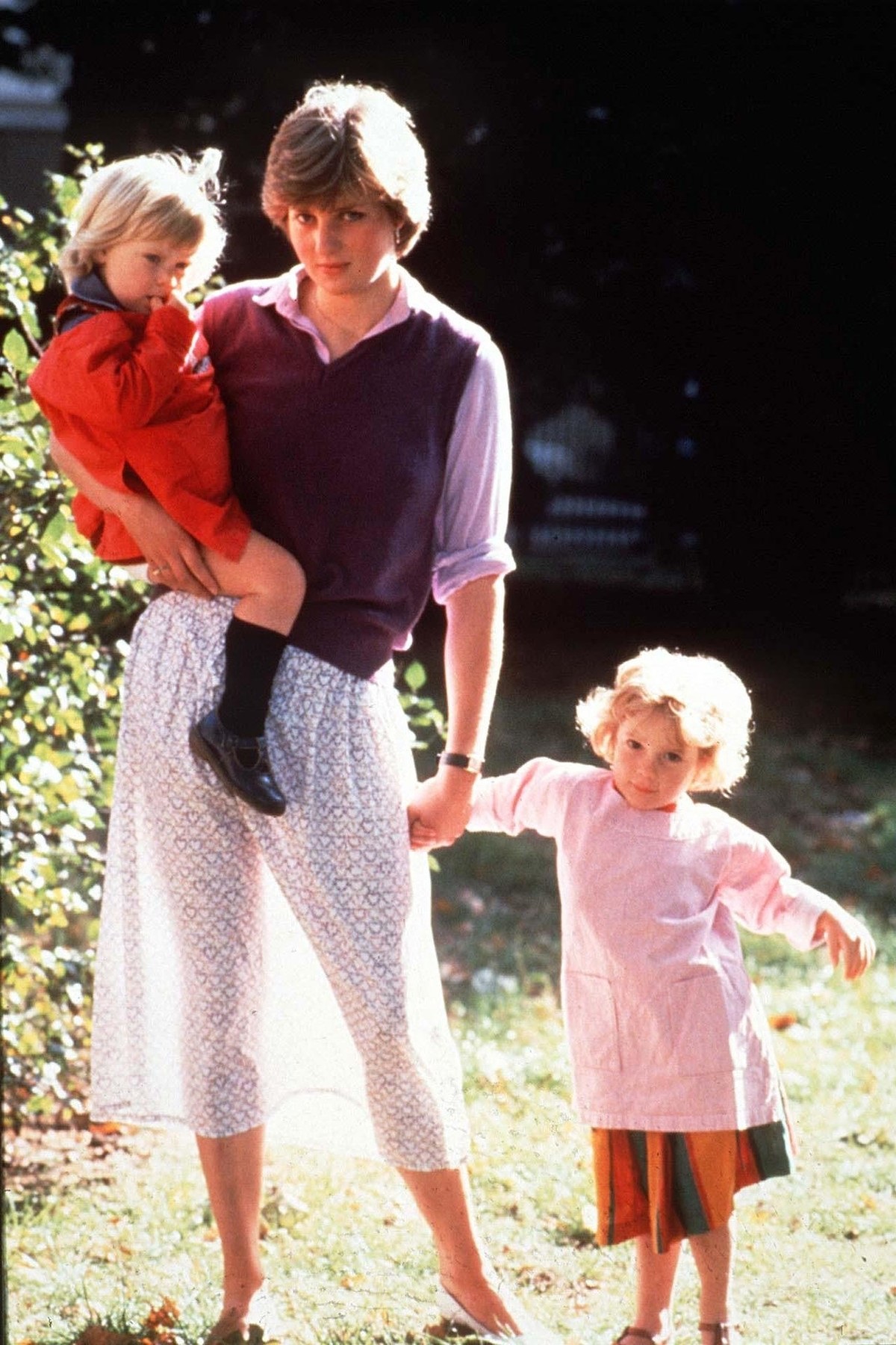 Lady Di într-o fustă cu buline și o vestă mov în timp ce ține o fetiță de mână și a alta în brațe în timp ce lucra la o școală de asistente în 1980, fiind unul dintre acele momente emblematice ale Prințesei Diana