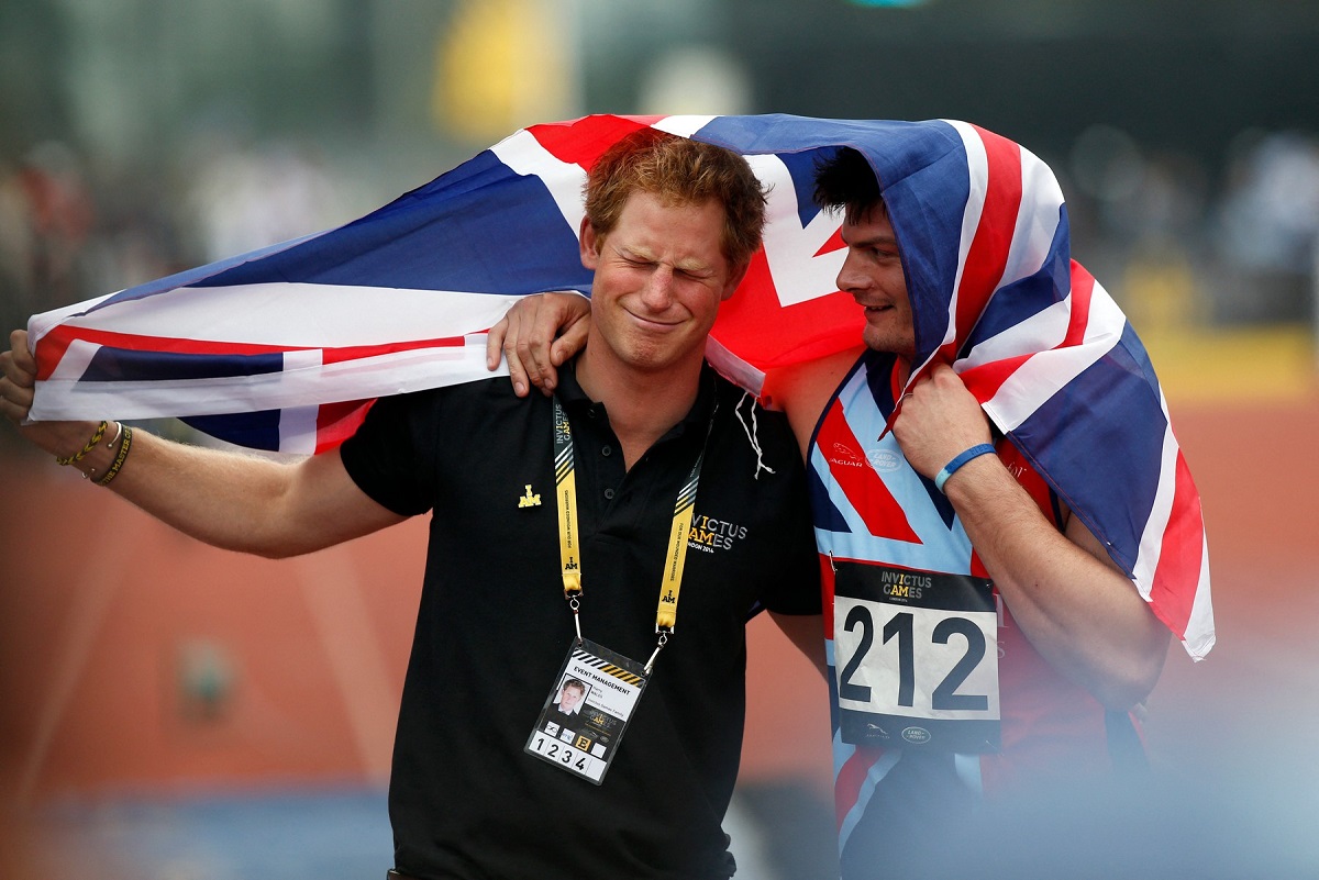 Prințul Harry alături de David Henson în timp ce se țin după umeri și sunt acoperiți cu steagul Marii Britanii pentru a celebra câștigul lui David la Jocurile Invictus din 2014