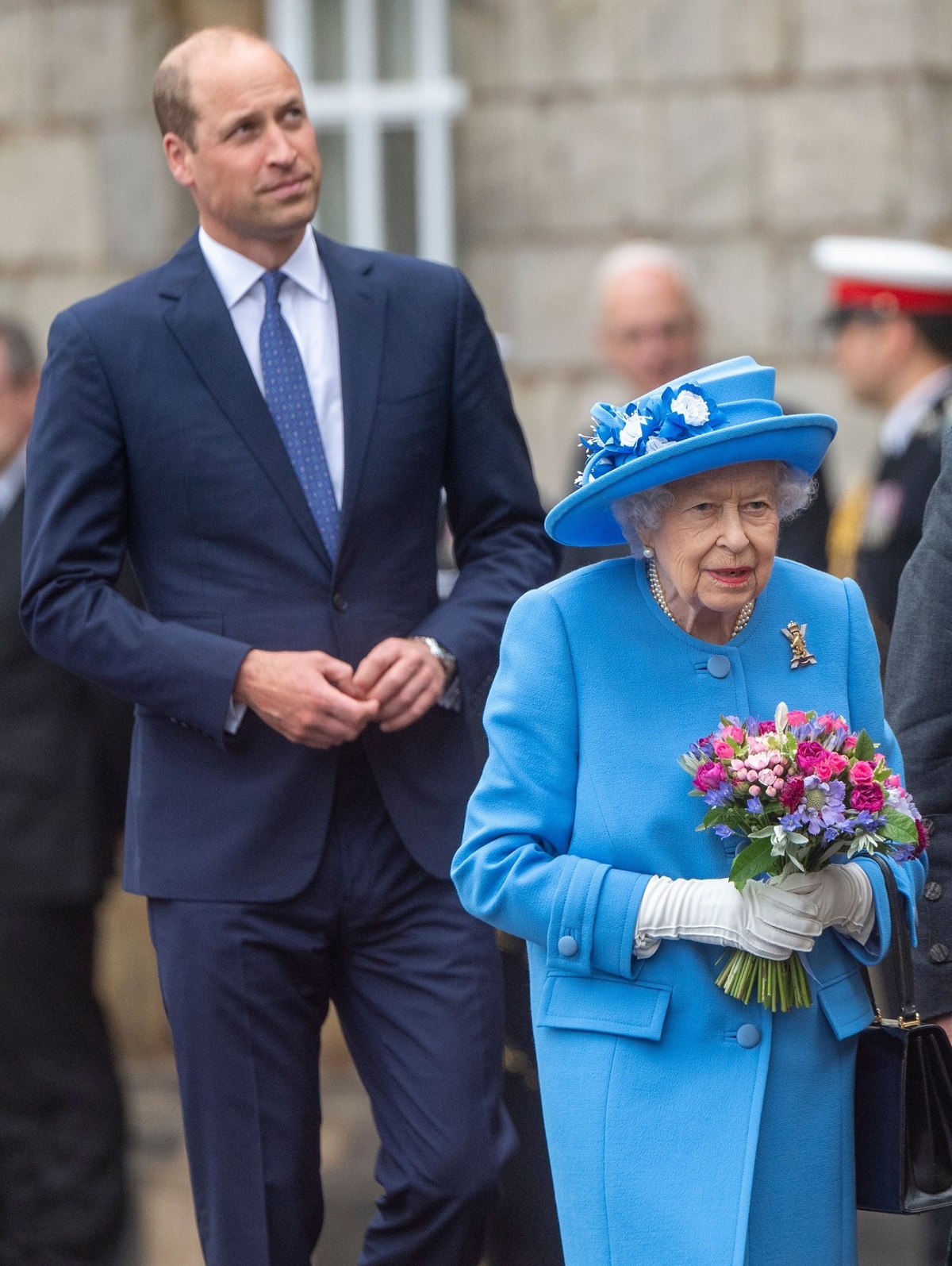 Prințul William îmbrăcat la costum în timp ce stă în spatele bunicii sale, Regina Elisabeta, în timpul unei vizite oficiale din Scoția