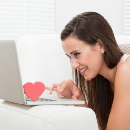 O femeie tânără folosește laptopul, în timp ce zâmbește. Tastatura laptopului are amplasată o inimioară în apropierea ecranului