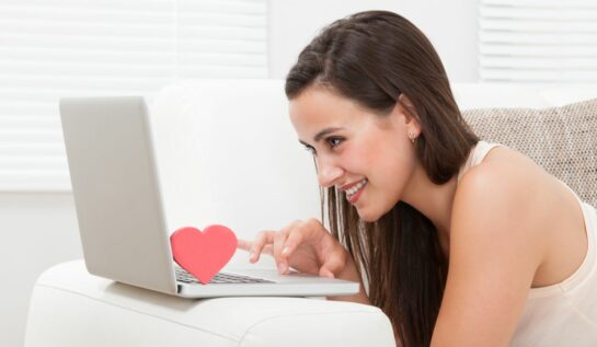 O femeie tânără folosește laptopul, în timp ce zâmbește. Tastatura laptopului are amplasată o inimioară în apropierea ecranului